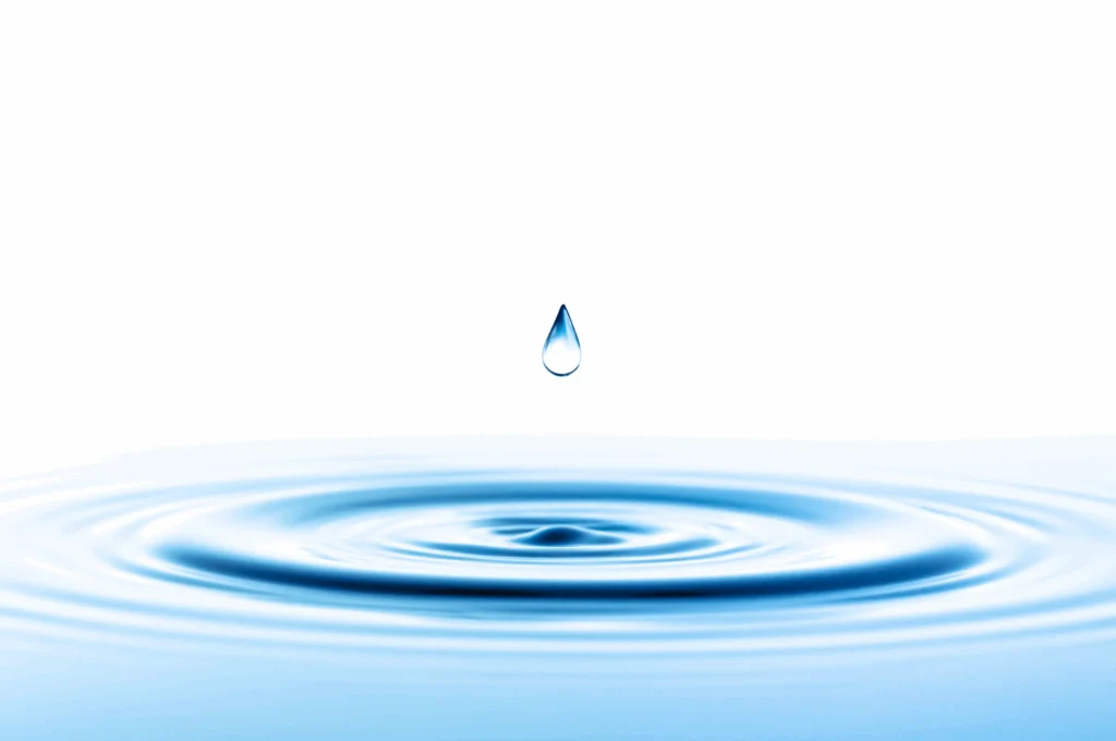Single water droplet falling towards concentric ripples on a clear blue water surface